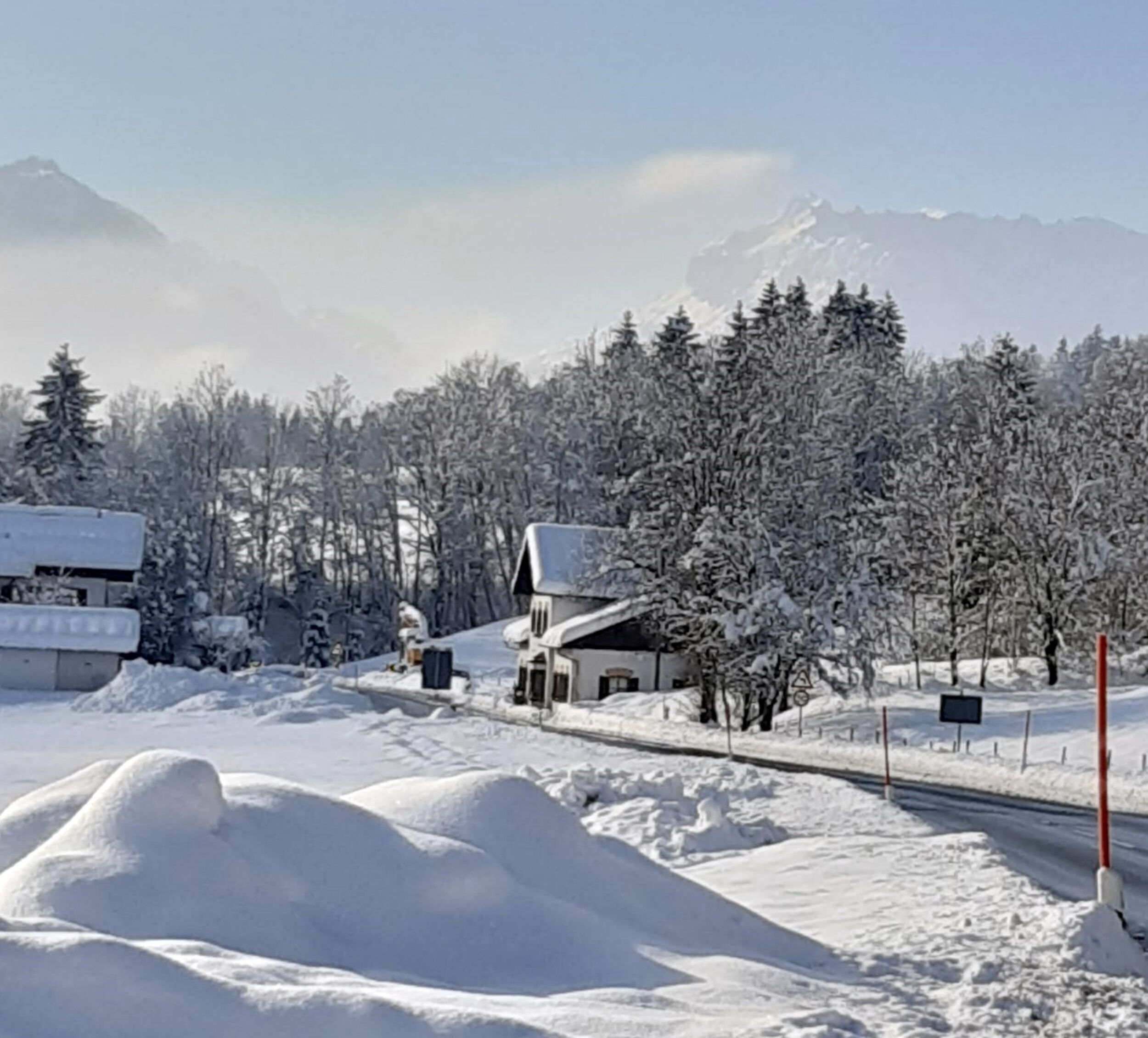 A snow-covered landscape with a road, a building with a snow-covered roof, and a mountain in the background.