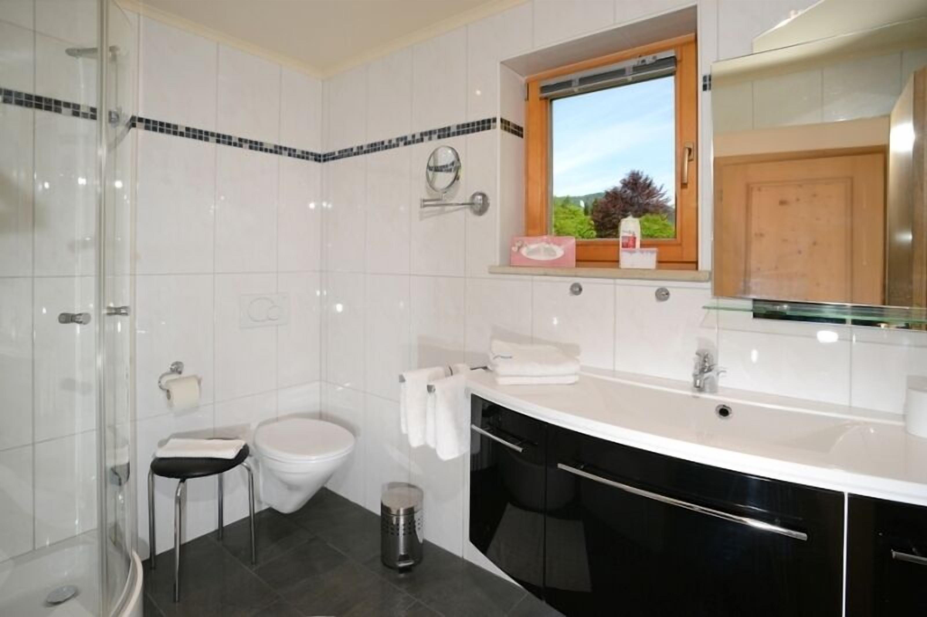 Un baño moderno con cabina de ducha de vidrio, inodoro, lavabo con tocador y ventana con vistas a la vegetación.