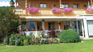 Een houten chalet met een balkon versierd met roze bloemen, een overdekt terras met buitenmeubels en een verzorgde tuin.
