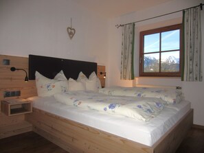 Un dormitorio con cama de madera, ropa de cama floral, una mesita de noche, una lámpara de pared y una ventana con vistas a montañas.