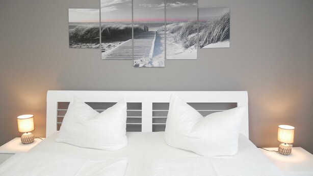 A hotel room with a large bed, two bedside lamps, and a wall-mounted artwork depicting a coastal scene. - Vacation apartment by the Baltic Sea - Apartment Kapitän 3 (Zinnowitz)