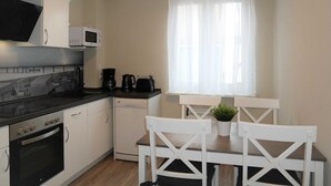 A compact kitchen with white cabinets, a black countertop, a microwave, a coffee maker, and a dishwasher. There is a window with sheer curtains and a small potted plant on the table. - Vacation apartment by the Baltic Sea - Apartment Kapitän 3 (Zinnowitz)