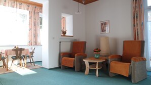Un salon chaleureux avec un plafond en bois, un coin repas avec une table et des chaises, et deux fauteuils ornés d’un vase de fleurs.