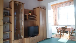 Une pièce comprenant un meuble en bois avec des portes en verre, un téléviseur posé sur un meuble, une table à manger avec des chaises et une fenêtre avec des rideaux.
