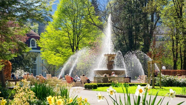 Un parc avec une fontaine, des bancs, et des gens assis ou en train de se promener.