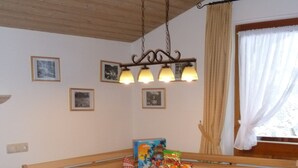A dining area with a wooden table, chairs, and a bench. There are framed pictures on the wall, a hanging light fixture, and a window with curtains. - Three-room vacation apartment, 40m², bedroom, children's room, living room, kitchenette, balcony (Reit im Winkl)