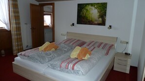 A double bed with patterned bedding, a wooden headboard, a nightstand with a lamp, and a painting of a forest on the wall. - Three-room vacation apartment, 40m², bedroom, children's room, living room, kitchenette, balcony (Reit im Winkl)