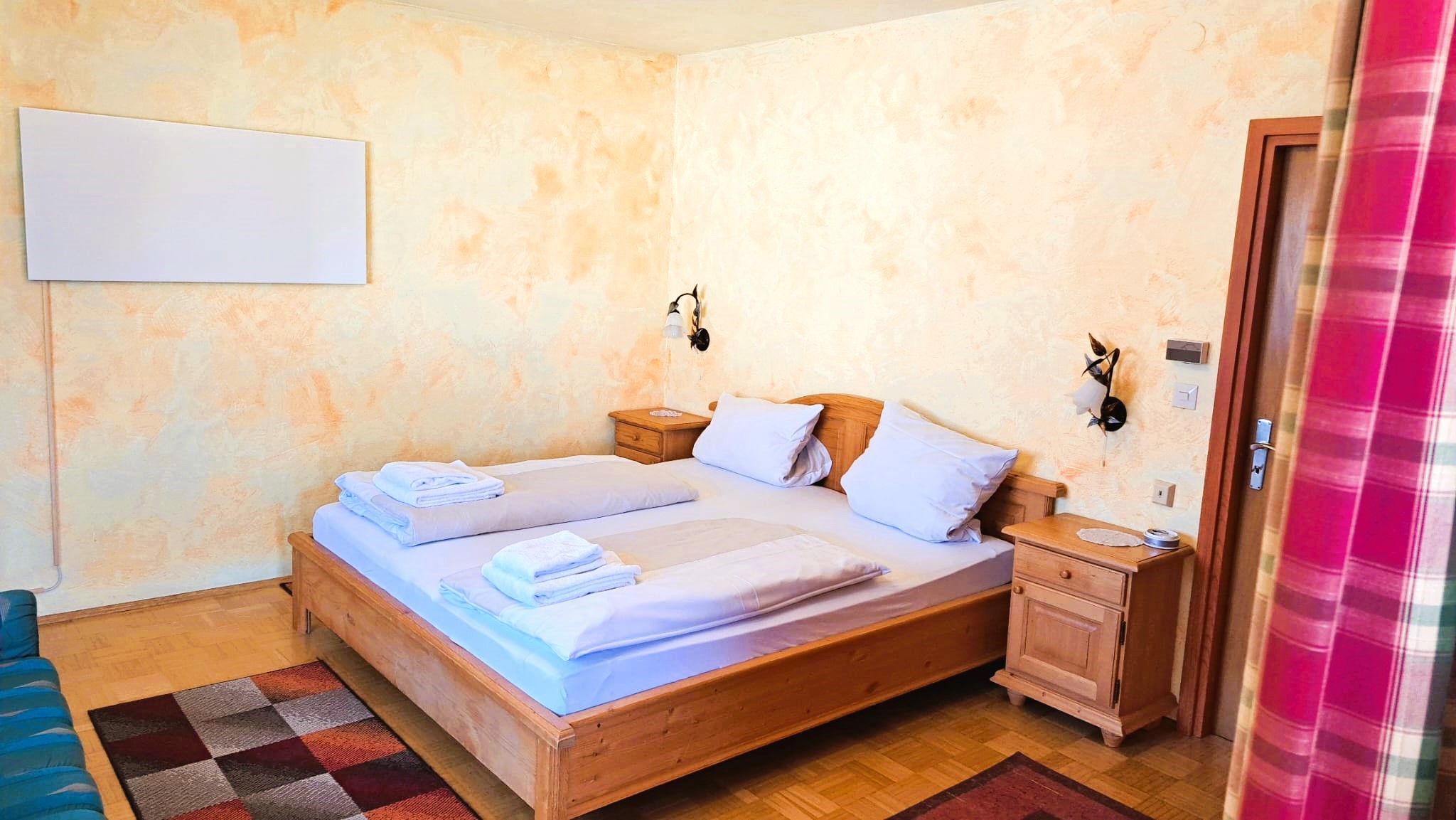 A bedroom with a wooden bed, two bedside tables, a nightstand, a wall-mounted light, and a checkered rug.
