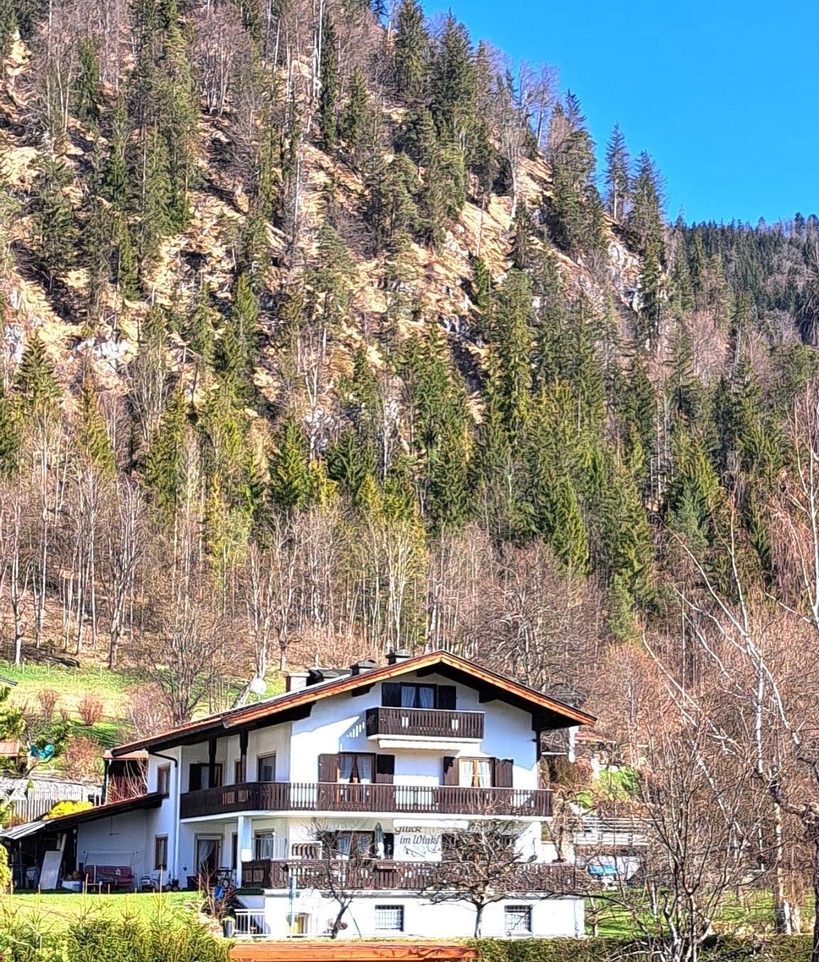 A two-story chalet with a balcony, surrounded by a forested hillside.