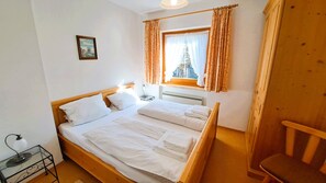 Una cama doble con ropa blanca, una mesita de noche con lámpara, una ventana con cortinas y una silla de madera.