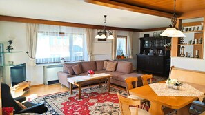 A living room with a sofa, a coffee table, a dining table with chairs, a bookshelf, and a large window with curtains.