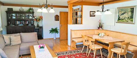 A living room with a sofa, a coffee table with a vase of flowers, a dining area with a bench and chairs, and a wooden cabinet with books and decorative items.