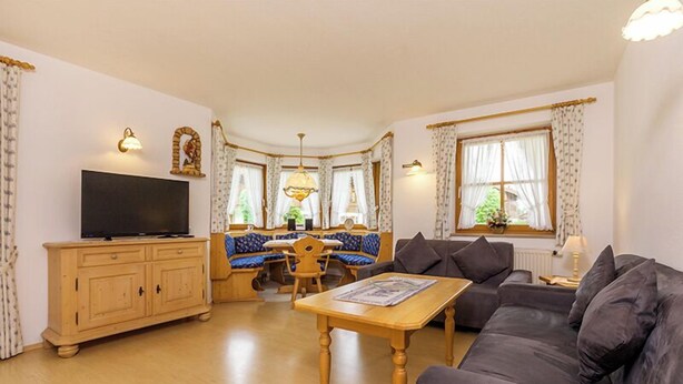 Una sala de estar con sofá, mesa de madera, televisión, zona de comedor con asientos azules y una ventana con cortinas.