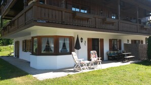 Un chalet de dos pisos con un balcón, un porche cubierto con mobiliario y vistas del paisaje circundante.