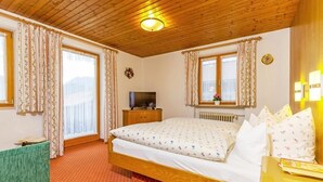 A bedroom with a wooden ceiling, a bed with patterned bedding, a television on a wooden cabinet, and a window with curtains. - Ferienwohnung (5) (Reit im Winkl)