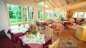 A dining area with multiple tables set for breakfast, featuring a variety of breakfast items and a large window offering a view of greenery outside. - 10 Apartment type D 71 sqm, balcony, 2 bedrooms, living room, max 4 p, WLAN (Ruhpolding)