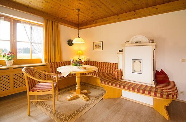 Een knus vertrek met een houten plafond, een ronde tafel met stoelen, een bank met kussens en een raam met gordijnen.