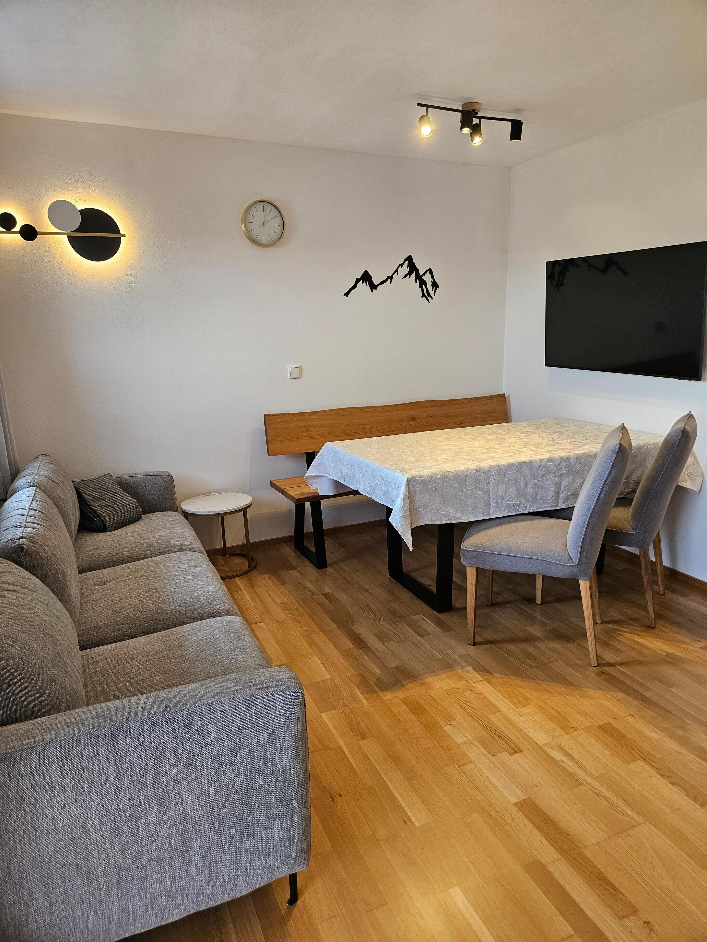 A cozy living room with a sofa, a small round table, a wooden bench, and a dining table with chairs. There is a wall-mounted clock and a flat-screen TV.