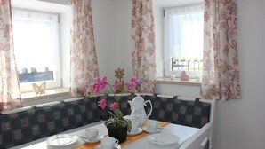 Un coin repas avec une table mise pour le thé, un vase de fleurs et une vue à travers une fenêtre ornée de rideaux.