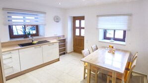A kitchen with a sink, cabinets, and a dining table with chairs. There is a window with blinds, a door, and a clock on the wall.