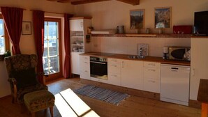 Una cocina con vigas de madera, armarios blancos, estufa, lavavajillas y una ventana con cortinas rojas.