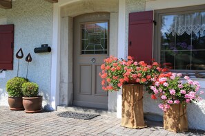 Welcome to our charming entrance, featuring a beautiful door and window above, surrounded by lush greenery and vibrant flowers.
