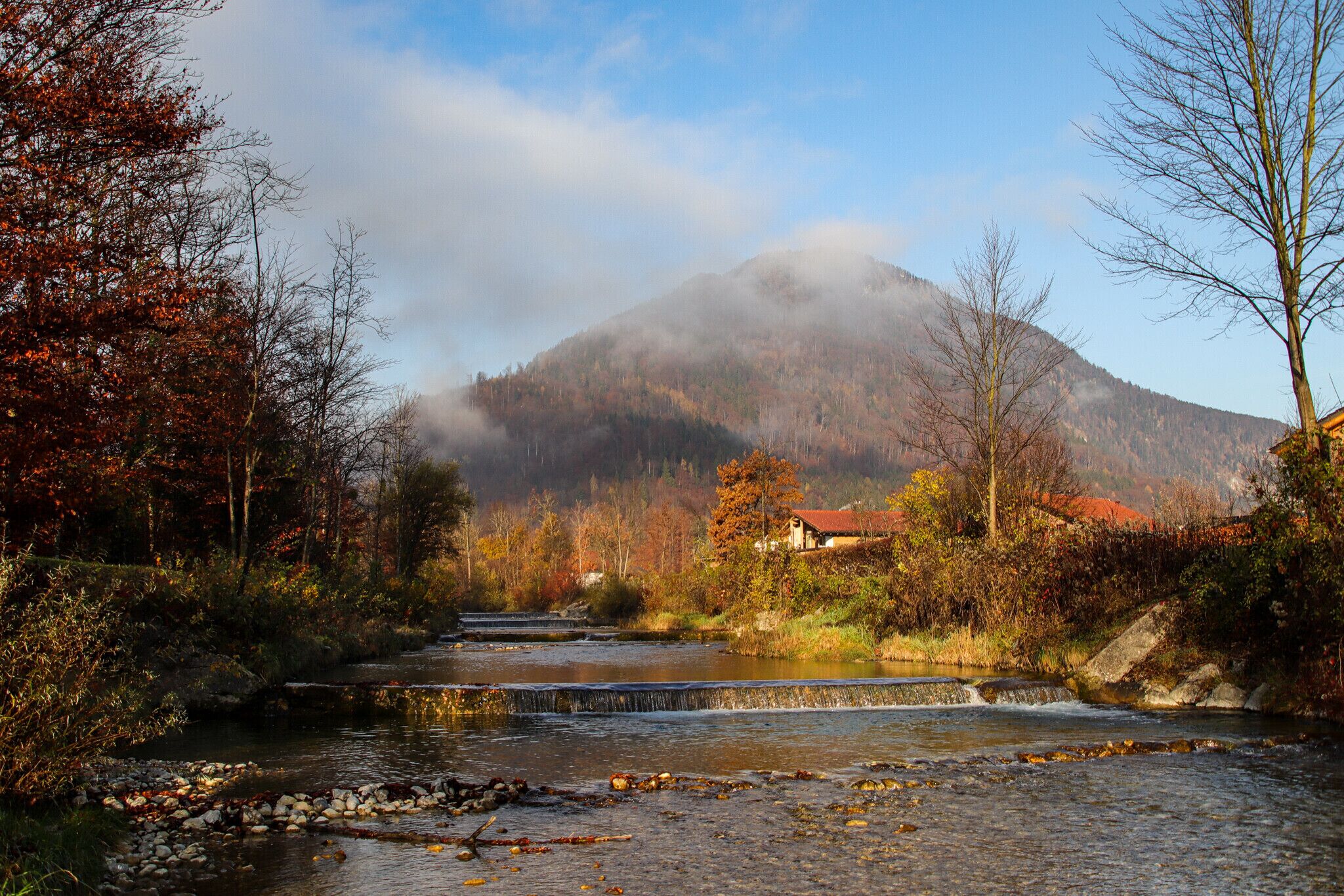 Une rivière qui traverse une vallée, avec une montagne en arrière-plan et une maison avec un toit rouge.