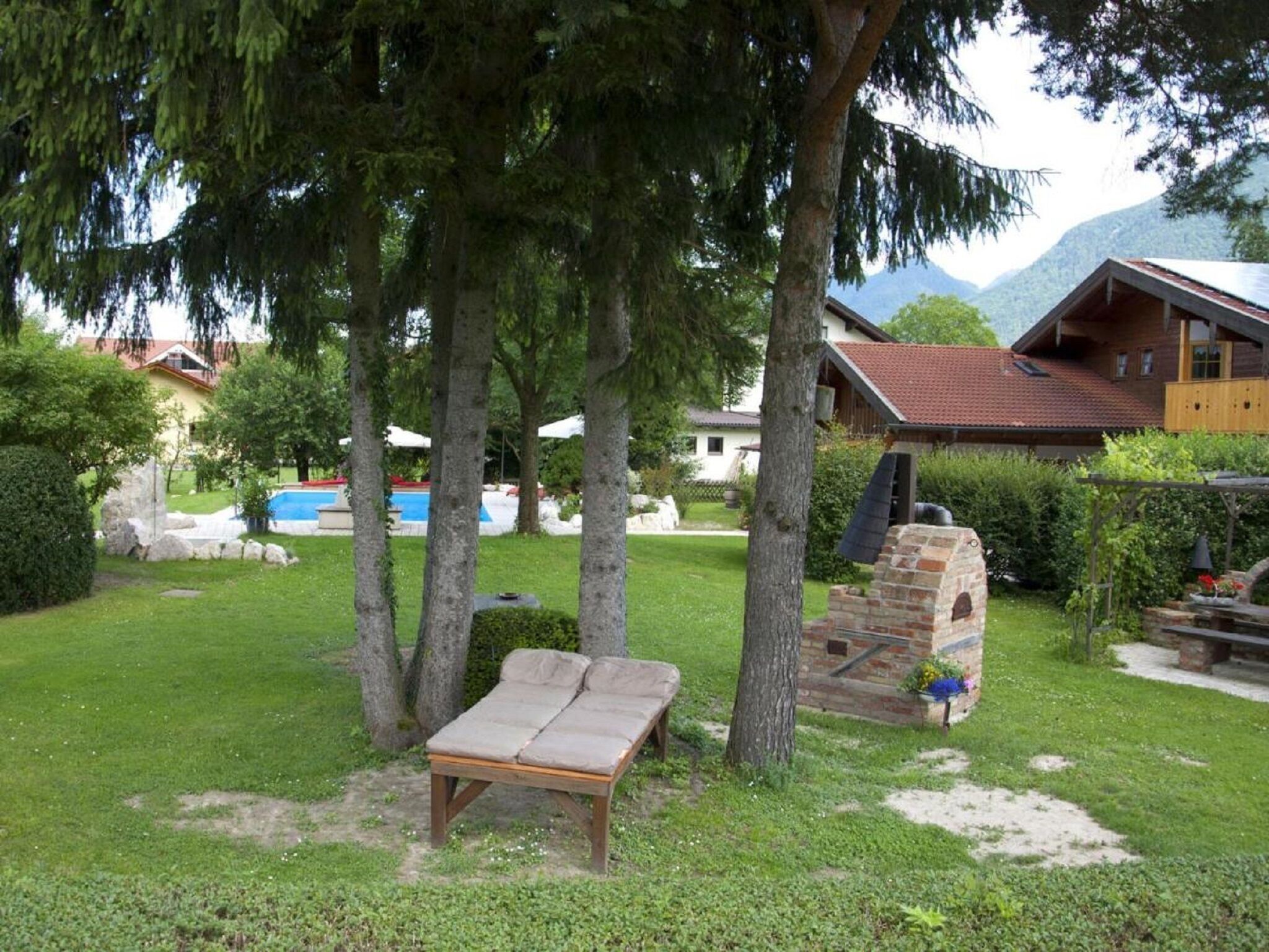 Eine hölzerne Bank unter einem Baum, ein Backofen und ein Schwimmbecken in einem Garten mit Häusern im Hintergrund.