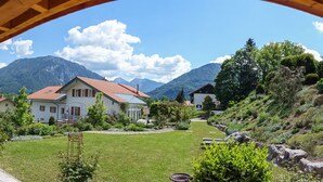 A view of a house with a red roof and white walls, surrounded by a well-maintained garden and mountains in the background. - Jasmin vacation apartment with south-facing terrace and mountain view (Ruhpolding)