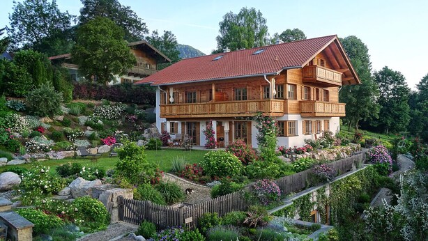 A large wooden house with a red-tiled roof, surrounded by a well-maintained garden with various plants and flowers. - Jasmin vacation apartment with south-facing terrace and mountain view (Ruhpolding)