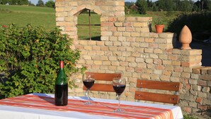 Una mesa con una botella y dos copas de vino tinto, un muro de ladrillo con una escultura de pájaro, y un banco.