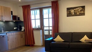 Ein modernes Wohnzimmer mit einer schwarzen Couch, einem Esstisch mit weißer Tischdecke und einer Küchenzeile mit Holzschränken und einem Fenster mit roten Vorhängen.