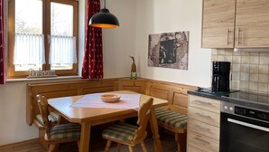 A cozy dining area with a wooden table and chairs, a pendant light, a window with curtains, a kitchen with wooden cabinets, and a coffee maker. - Apartment no. 4, 1-2 persons, 60 sqm, combined living room/bedroom, terrace (Schönau am Königssee)