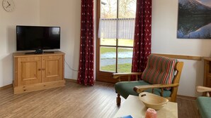 A living room with a television on a wooden cabinet, a striped armchair, a table with a bowl and a candle, and a window with red curtains. - Apartment no. 4, 1-2 persons, 60 sqm, combined living room/bedroom, terrace (Schönau am Königssee)