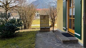A stone pathway leading to a yellow building with green shutters, a wooden fence, and a view of mountains in the background. - Apartment no. 4, 1-2 persons, 60 sqm, combined living room/bedroom, terrace (Schönau am Königssee)