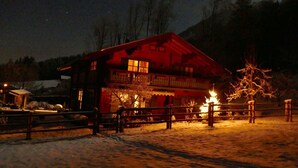 A snow-covered landscape with a red wooden building featuring a balcony and a lit tree in the foreground. - Ferienwohnung 2 bis 4 Personen, 60 qm, 2 Schlafzimmer, Balkon (Schönau am Königssee)