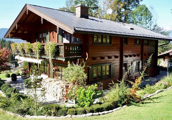 A wooden cabin with a balcony, surrounded by a garden with various plants and a stone wall.