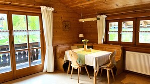 A wooden cabin with a dining area featuring a table and chairs, large windows with curtains, and a view of greenery outside. - Ferienwohnung 2 bis 4 Personen, 60 qm, 2 Schlafzimmer, Balkon (Schönau am Königssee)
