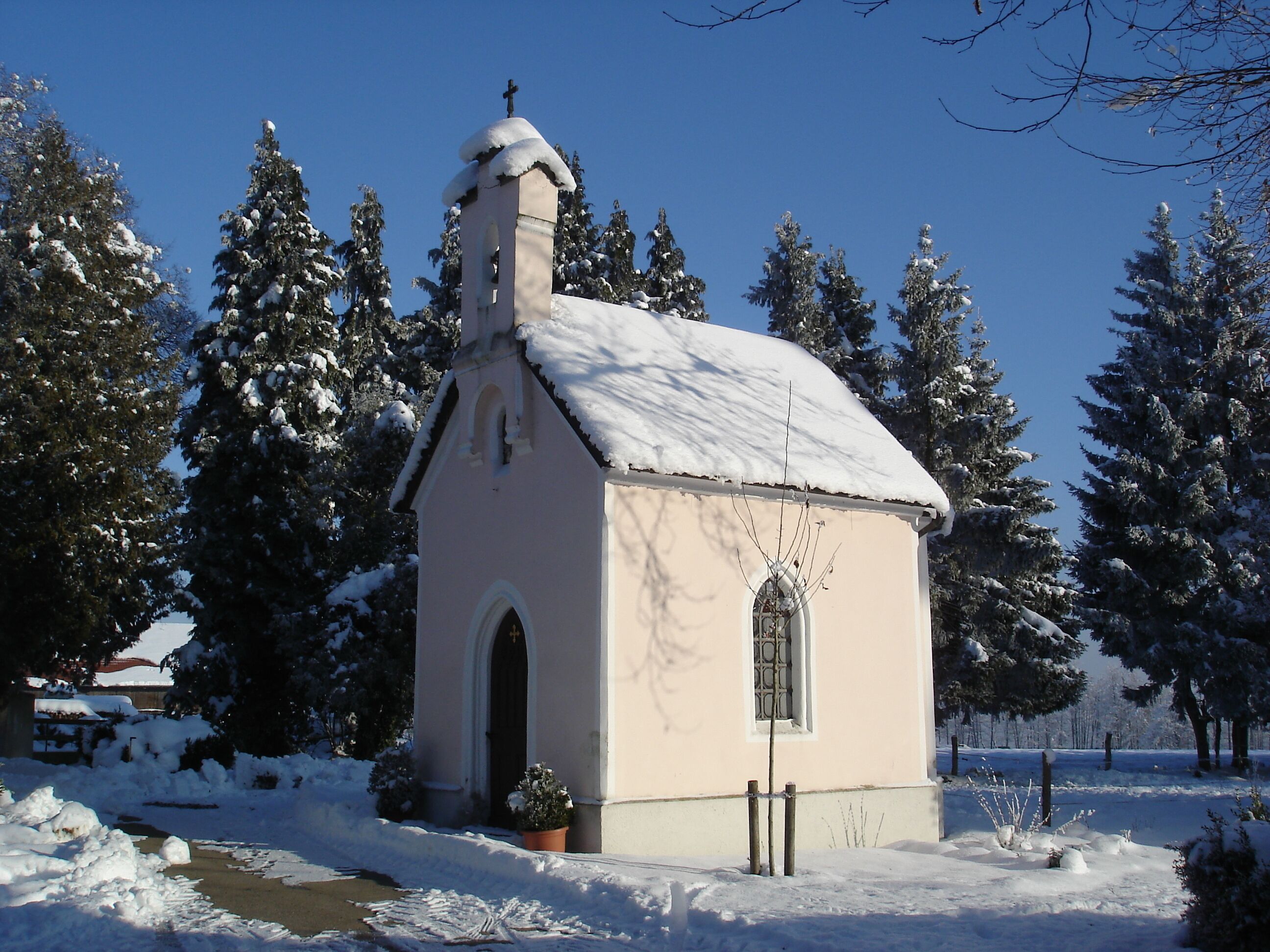 Una piccola chiesa con tetto imbiancato dalla neve e una croce sulla sommità, circondata da alberi innevati.