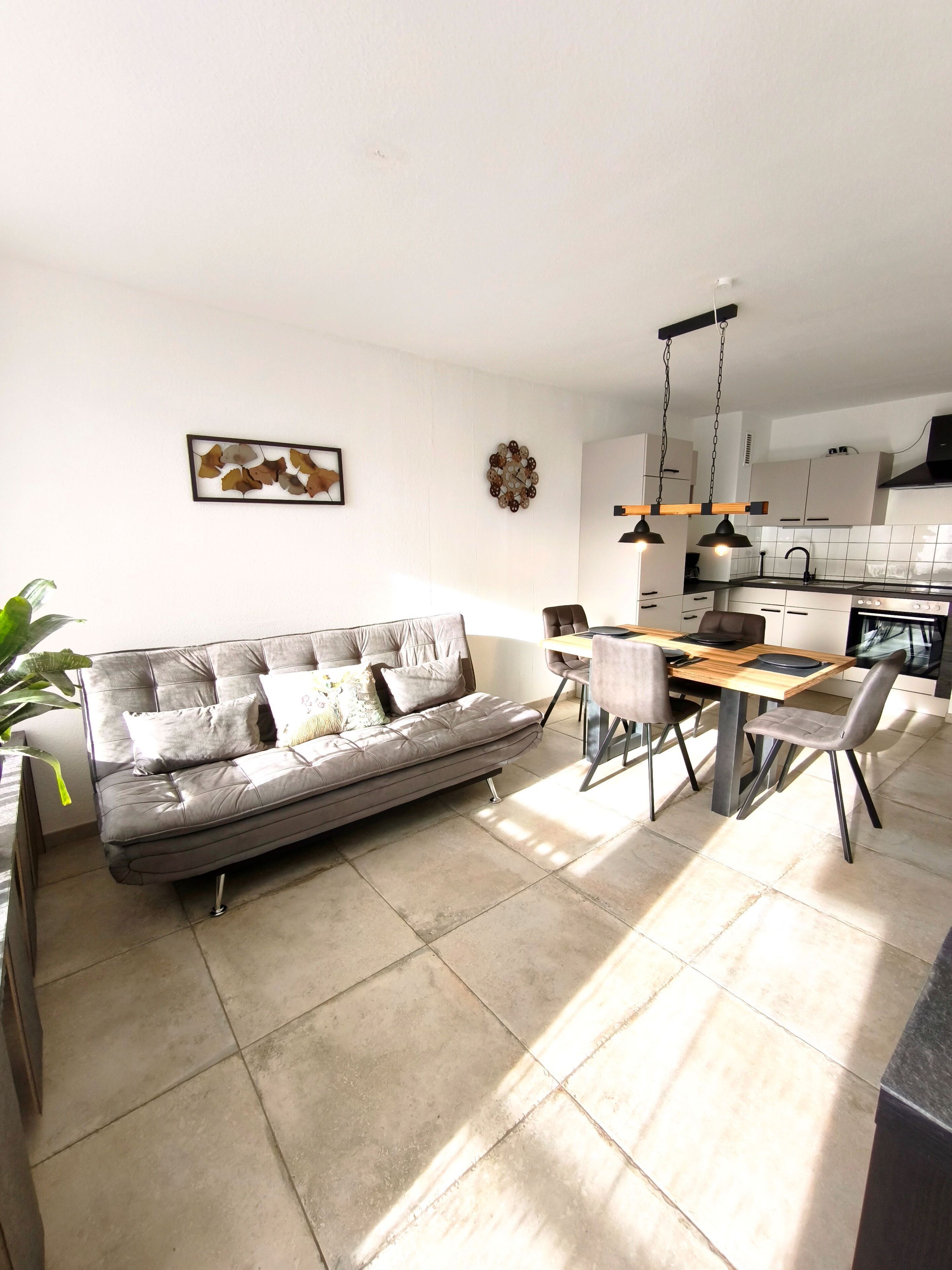 Una sala de estar moderna con un sofá gris, una mesa de comedor con sillas, una zona de cocina con armarios y una estufa, y un reloj fijado a la pared.