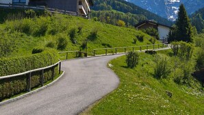 A modern building with a balcony, surrounded by lush greenery and a winding road, with mountains in the background.