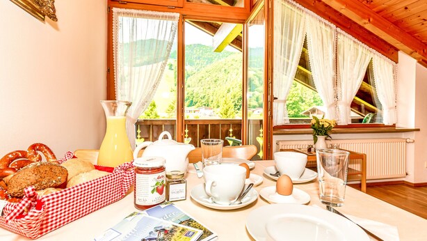 Une table de petit-déjeuner dressée avec du pain, des viennoiseries, une carafe de jus de fruits, une tasse, un verre et un livre.