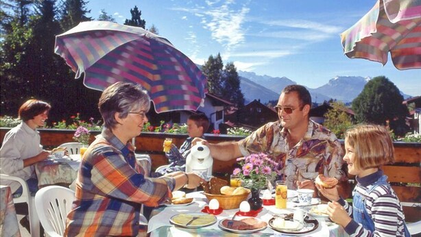 A group of people dining outdoors at a table with a colorful tablecloth, surrounded by chairs and umbrellas. - (8) Zwei-Raum-Ferienwohnung 41qm, Dusche/WC, Extra-Schlafraum, Küche, 2 Balkone (Reit im Winkl)