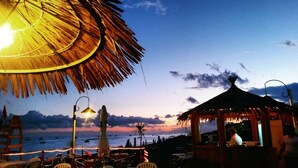 En restaurang vid stranden med ett halmtak, uteservering och utsikt över havet vid solnedgången.