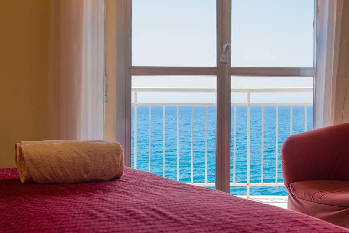 Een kamer met een bed, een rode stoel en uitzicht op de zee door een raam.