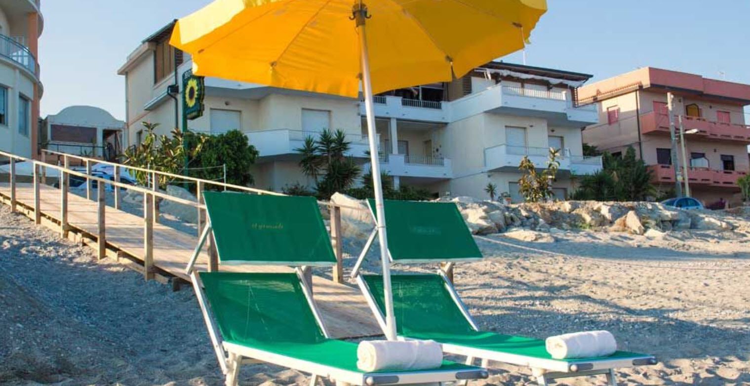 A beach with two green lounge chairs under a yellow umbrella, a wooden walkway, and buildings in the background.