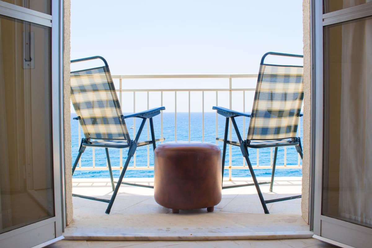 Een balkon met twee geruite stoelen en een klein voetenbankje, met uitzicht op een wateroppervlak.