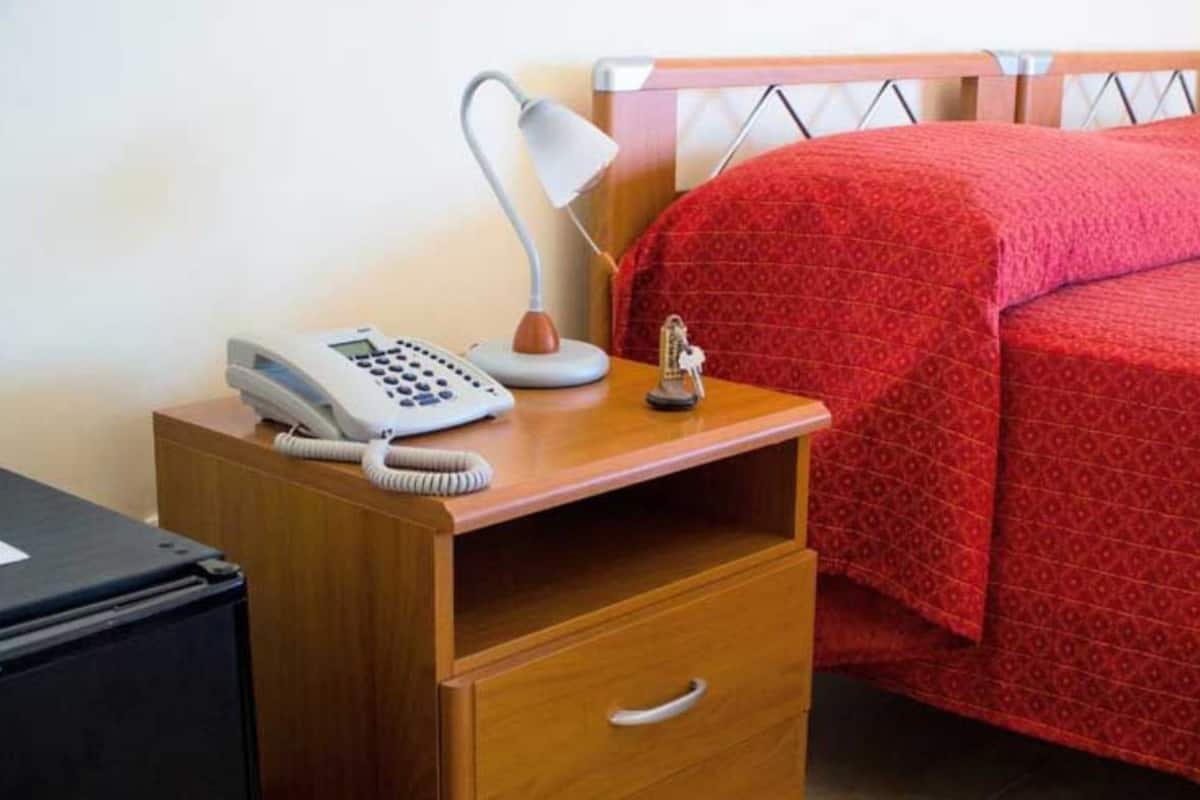 Een net opgemaakt bed met een rode deken, een bedlampje en een telefoon op een nachtkastje van hout.