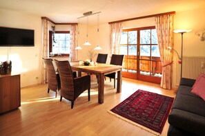 Un coin repas avec une table et des chaises en bois, un téléviseur fixé au mur et une grande fenêtre avec des rideaux.