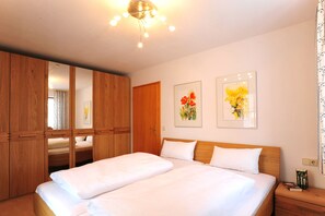 A hotel room with a bed, wooden wardrobe, two framed floral paintings, and a nightstand with books.